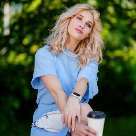 Young beautiful blonde girl in a blue short dress and with a white belt bag walks in a green park. He sits on a bench and drinks takeaway coffee.